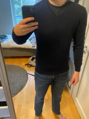 Mörkblå tröja i merinoull - Snygg mörkblå tröja med V-ringning och ljusgrå armbågslappar. Tröjan har en smal passform och långa ärmar, perfekt för en stilren och enkel look. Passar till både jeans och chinos för en avslappnad men ändå uppklädd stil. Väldigt fint skick!