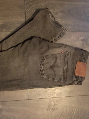 Svarta Levi's jeans med läderpatch - Säljer ett par svarta Levi's jeans med klassisk femficksdesign och brun läderpatch bak. Jeansen har raka ben och är tillverkade i slitstark denim. Perfekt för dig som gillar en tidlös och cool look.