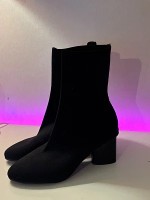 Svarta stretchiga stövletter med klack - Säljer ett par svarta stövletter/ sock stövlar med stretchigt tyg och fyrkantig tå. Skorna har en hög, bred klack och dragkedja på insidan. Aldrig använda