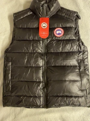 Svart dunväst från Canada Goose - Säljer en svart dunväst från Canada Goose med klassisk logopatch på bröstet. Västen har hög krage, dragkedja framtill och två stora fickor. Materialet är glansigt och quiltat, perfekt för kyliga dagar.