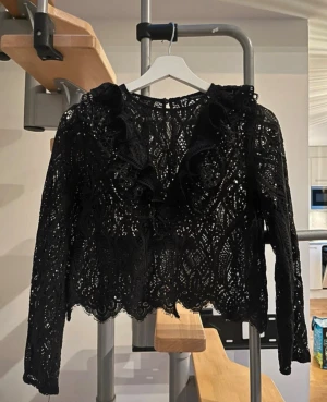 Svart spetsblus med volanger - Snygg svart blus i spets med långa ärmar och dekorativa volanger framtill. Blusen har ett genombrutet mönster och vågig kant nertill, vilket ger en trendig och elegant look. Perfekt att styla med jeans eller kjol för en cool outfit.