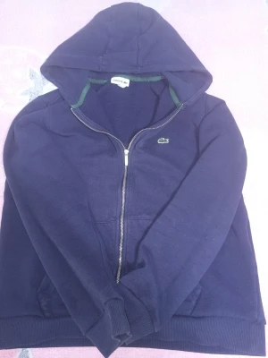 Mörkblå hoodie från Lacoste - Snygg mörkblå hoodie från Lacoste med dragkedja och klassisk krokodillogga på bröstet. Hoodien har huva, ribbade muddar och gröna detaljer vid kragen. Perfekt för en avslappnad och sportig stil.