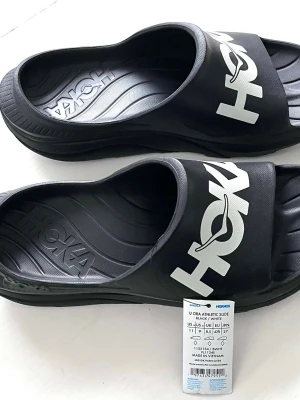 Svarta slides från Hoka - Säljer ett par svarta slides från Hoka med stor vit logga över foten. Sandalerna har en sportig look och är tillverkade i syntetmaterial med formad sula för extra komfort. Perfekta för chill dagar eller efter träningen.Skickar endast med PostNord.Vänligen observera att jag inte har öppetköp.