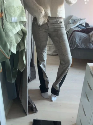 Grå jeans med svarta detaljer - Säljer ett par grå jeans med svarta paneler längs benen. Jeansen har en rak passform och normal midja. De har klassisk femficksdesign och stängs med dragkedja och knapp. Perfekta för dig som vill ha en unik twist på dina vardagsjeans. Dom är egensydda med det svarta!