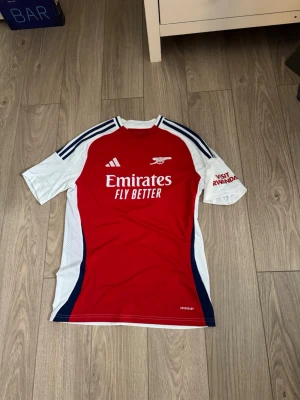 Arsenal matchtröja Adidas röd/vit - Snygg Arsenal matchtröja från Adidas i rött och vitt med blå detaljer. Tröjan har klubbens logga och sponsortryck framtill samt 'Visit Rwanda' på ärmen. Tillverkad i lätt och ventilerande funktionsmaterial, perfekt för fotboll eller träning. Aldrig använd så inga hål märken eller slitage. Levereras alltid väl förpackat samt snabbt! Storlek är 170 så passar xs/s. Mvh! Sökord: adidas,arsenal,fotboll. 