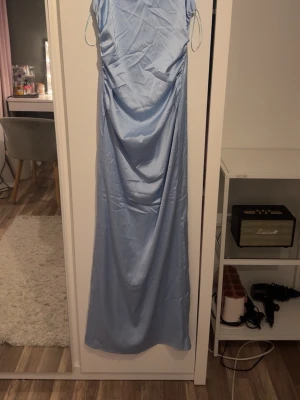 Ljusblå balklänning  - Säljer en elegant ljusblå långklänning med tunna axelband och dragkedja i ryggen, men den syns knappt. Klänningen har en mjukt draperad framsida och är tillverkad i ett glansigt material som ger en lyxig känsla. Perfekt för sommarens alla tillfällen.