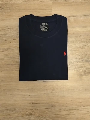 Mörkblå t-shirt från Polo Ralph Lauren - Klassisk mörkblå t-shirt från Polo Ralph Lauren med rund halsringning och den ikoniska röda logotypen broderad på bröstet. Tillverkad i mjuk bomull och har en normal passform. Perfekt basplagg för en stilren look.