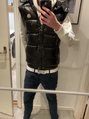 Svart väst från Moncler - Snygg svart dunväst från Moncler med glansig finish och huva. Västen har dragkedja framtill, två sidofickor med dragkedja och Moncler-logga på bröstet. Perfekt för lager-på-lager och ger en clean streetwear-look.