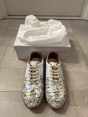Vita sneakers med färgstänk från Maison Margiela - Unika vita sneakers från Maison Margiela med färgstänk i blått, gult och svart. Skorna har låga skaft, beige snörning och bruna detaljer på insidan samt gummisula. Tillverkade i läder och textil, perfekta för dig som vill sticka ut med din stil.