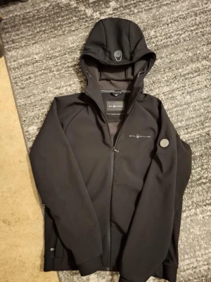 Svart vindjacka från Sail Racing - Stilren svart vindjacka från Sail Racing med huva och hel dragkedja framtill. Jackan har diskreta logotyper på bröstet och ärmen samt praktiska fickor. Tillverkad i ett mjukt och följsamt material som passar perfekt för blåsiga dagar.