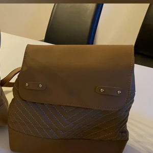 Brun ryggsäck i konstläder - Snygg brun ryggsäck i konstläder med dekorativa sömmar och lock med två knappar. Justerbara axelband och praktisk storlek för vardagsbruk. Enkel men trendig design som passar till många stilar.