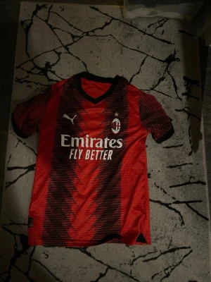 AC Milan matchtröja Puma röd/svart - Säljer en AC Milan matchtröja från Puma i rött och svart med klubbmärke och sponsortryck på bröstet. Tröjan har korta ärmar, V-ringning och är tillverkad i ett lätt, ventilerande material perfekt för fotboll eller träning.