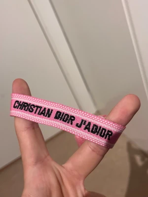 Christian Dior J'Adior rosa armband - Snyggt rosa tygarmband från Christian Dior med texten 'J'Adior' i svart. Armbandet har en vävd struktur och är perfekt för dig som vill ha en trendig accessoar med lyxig känsla. Justerbar storlek och cool design.