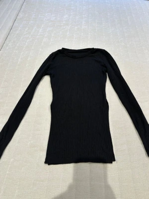 Svart ribbad långärmad topp - En svart, figurnära långärmad topp med ribbad struktur och rund halsringning. Perfekt basplagg som är lätt att matcha med olika outfits. Materialet ser ut att vara stretchigt och mjukt för en bekväm passform.