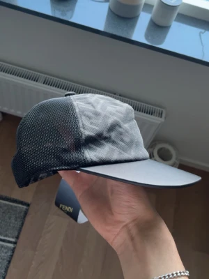 Svart och grå keps från Fendi - Snygg keps från Fendi i svart och grått med diskret FF-mönster på framsidan. Baksidan är i mesh för extra ventilation och justerbar rem med guldfärgad Fendi-detalj. Perfekt accessoar för en trendig streetstyle-look.