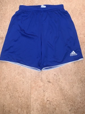 Blå sportshorts från Adidas - Säljer ett par blå sportiga shorts från Adidas med vit logga på vänster ben. Shortsen har resår i midjan och är tillverkade i ett lätt och snabbtorkande material, perfekta för träning eller chill. Klassisk passform och stilren design.