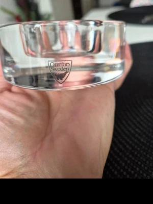 Glasljusstake Orrefors Sweden - Säljer en rund, transparent glasljusstake från Orrefors Sweden. Ljusstaken har en stilren och enkel design med Orrefors-loggan ingraverad på sidan. Perfekt som inredningsdetalj för att skapa mysig stämning hemma.