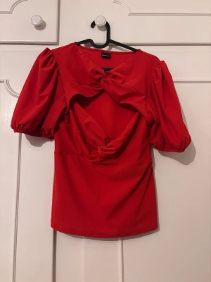 Röd topp med puffärm och rosetter - Röd topp med puffärmar och två stora rosetter framtill. Toppen har en öppen detalj vid bröstet. Perfekt för dig som vill sticka ut med en unik och färgstark look.