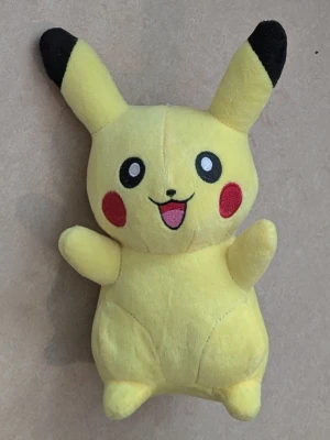 Pikachu mjukisdocka  - Gul mjukisdocka föreställande Pikachu från Pokémon.  Dockan har svarta öronspetsar, röda kinder och ett glatt ansikte. Tillverkad i mjukt plyschmaterial. Längd ca 23 cm