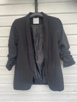 Svart kavaj från Pieces - Stilren svart kavaj från Pieces med klassisk krage och upprullade ärmar. Kavajen har en rak passform och är helfodrad med slätt tyg på insidan. Perfekt för en trendig och enkel look.