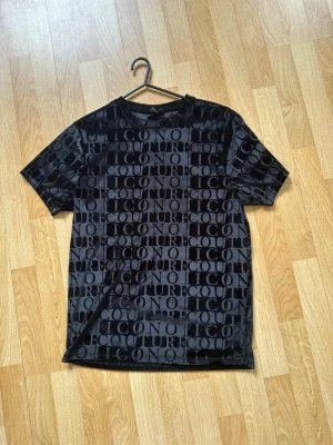 Svart t-shirt från Couture ICON - Snygg svart t-shirt från Couture ICON med stort, upprepat logotypmönster över hela plagget. T-shirten har klassisk passform, rund hals och korta ärmar. Materialet känns mjukt och har en lätt glansig finish som ger en exklusiv vibe.