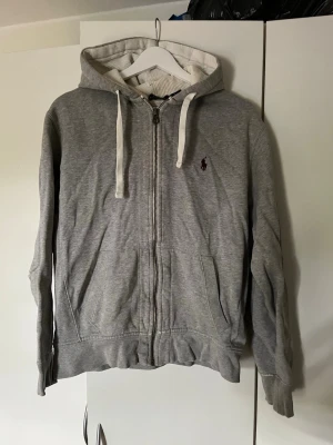 Grå zip hoodie från Polo Ralph Lauren - Snygg grå hoodie med dragkedja från Polo Ralph Lauren. Klassisk design med vit dragsko i huvan, två fickor framtill och broderad logga på bröstet. Tillverkad i mjuk bomullsblandning, perfekt för chill dagar eller när du vill ha en clean look.