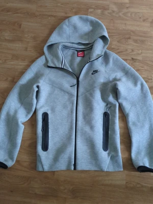 Grå hoodie från Nike med dragkedja - Snygg grå hoodie från Nike med svart logga på bröstet och hel dragkedja framtill. Två vertikala fickor med svarta dragkedjor och en stor huva. Tillverkad i mjukt material som känns bekvämt och sportigt.    Kod ord NIKE, KOFTA , NIKE TECH, HOODIE