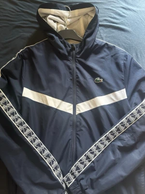 Marinblå vindjacka från Lacoste - Snygg marinblå vindjacka från Lacoste med vit diagonal rand över bröstet och vita mönstrade band längs ärmarna. Jackan har huva, dragkedja och meshfoder på insidan. Klassisk krokodillogga på bröstet och sportig look.