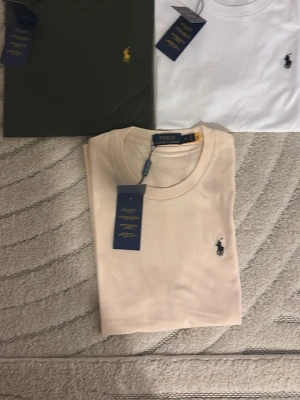Beige t-shirt från Polo Ralph Lauren - Snygg beige t-shirt från Polo Ralph Lauren med klassisk rund hals och ikoniska broderade loggan på bröstet. Tillverkad i mjuk bomull och har en clean, stilren look som passar till allt. Perfekt basplagg för en enkel och fräsch outfit.
