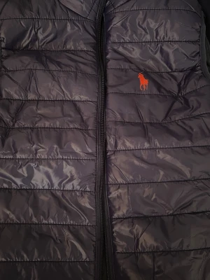 Svart dunväst från Polo Ralph Lauren - Snygg svart dunväst från Polo Ralph Lauren med klassisk röd logga på bröstet. Västen har horisontella sömmar, dragkedja framtill och är tillverkad i glansigt material. Perfekt för lager på lager och en stilren look.