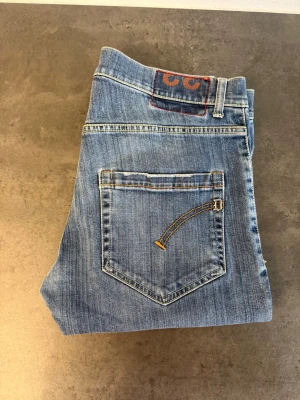 Blå jeans från Dondup W33 - Säljer ett par klassiska blå jeans från Dondup med raka ben och femficksmodell. Jeansen har snygga slitningar och kontrastsömmar, samt en broderad detalj på bakfickan. Tillverkade i bomull för en skön och avslappnad look.