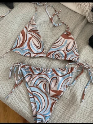 Brun och blå mönstrad bikini - Trendig bikini med swirl-mönster i brunt, blått och vitt. Triangeltopp med knyt i nacken och ryggen samt matchande trosa med knyt i sidorna. Perfekt för sommaren och strandhäng!