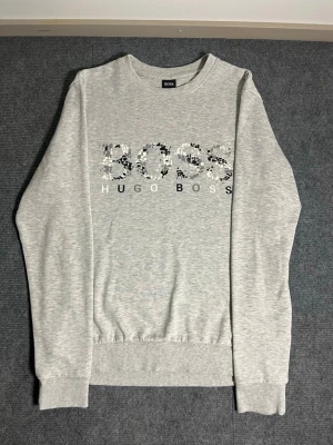 BOSS Hugo Boss – Light Grey Sweatshirt (M) - Modern and comfortable Hugo Boss sweatshirt in light grey. Size M, length 67 cm, width 50 cm – designed for a clean, contemporary fit. The front features the signature BOSS HUGO BOSS logo in a stylish pixel‑pattern print, giving the piece a subtle yet distinctive look.  ✅ Condition: Excellent – no stains, no damage 💯 Authenticity: 100% genuine Hugo Boss product 🧵 Material: Slightly thinner cotton blend – soft, breathable, and comfortable to wear; not too warm, making it ideal for everyday use 📦 
