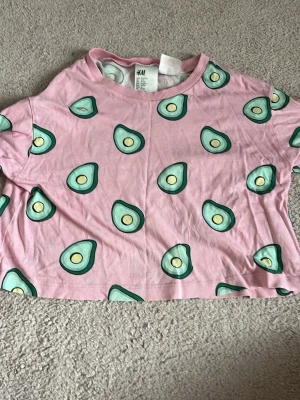 Rosa croppad t-shirt med avokadotryck - Söt rosa croppad t-shirt från H&M med ett lekfullt avokadotryck över hela plagget. T-shirten har korta ärmar och rund halsringning. Perfekt för dig som vill sticka ut med en färgglad och rolig stil.