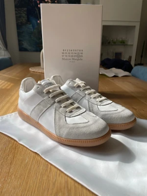 Maison Margiela vita och grå sneakers - Maison Margiela sneakers i vitt och ljusgrått med ovandel i slätt läder och mocka. Klassisk låg modell med beige gummisula och vita snören. Snygg minimalistisk design som passar till det mesta. Märkeslogga på plösen och insidan.