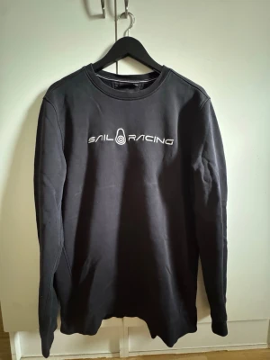 Svart sweatshirt från Sail Racing - Svart sweatshirt från Sail Racing med vitt broderat logotyptryck på bröstet. Tröjan har rund halsringning och långa ärmar. Tillverkad i mjukt material som känns skönt mot huden. Perfekt för en avslappnad och sportig stil.