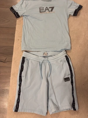 Ljusblått EA7 Emporio Armani set - Snyggt ljusblått set från EA7 Emporio Armani med t-shirt och shorts. T-shirten har logga framtill och svarta detaljer med EA7-tryck på ärmarna. Shortsen har dragsko i midjan, sidofickor och svarta loggband längs sidorna. Perfekt för en avslappnad och trendig look.