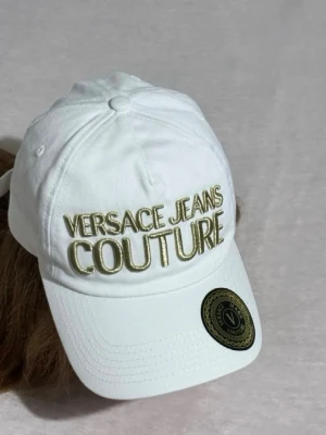Vit keps från Versace Jeans Couture - Snygg vit keps från Versace Jeans Couture med broderad logga i guld framtill. Klassisk böjd skärm och justerbar rem bak. Tillverkad i bomull för en bekväm och luftig känsla. Perfekt accessoar för att lyfta din outfit.     Pris kan diskuteras