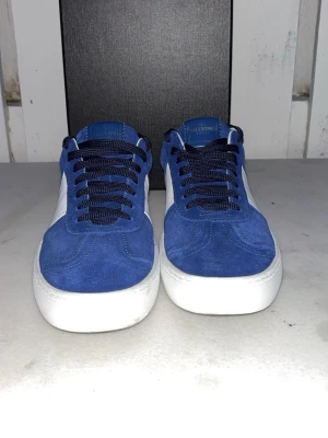 Valentino Flycrew  - Snygga blåa sneakers från Valentino med vit sula och vita detaljer på sidan. Ovandelen är i mocka och skorna har svarta skosnören. På hälen finns små nitar som ger en cool touch. Perfekta för dig som vill ha något stilrent men ändå sticker ut. Pris kan diskuteras