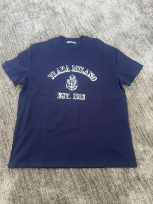 Mörkblå Prada Milano t-shirt - Snygg mörkblå t-shirt från Prada med vit text 'PRADA MILANO EST. 1913' och emblem på bröstet. Klassisk passform med rund hals och korta ärmar. Perfekt för dig som gillar exklusiva märken och stilren design.