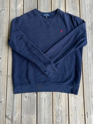 Mörkblå sweatshirt från Polo Ralph Lauren - Klassisk mörkblå sweatshirt från Polo Ralph Lauren med rund halsringning och röd broderad logga på bröstet. Tröjan har långa ärmar och ribbade muddar vid ärmslut och nederkant. Perfekt för en avslappnad och stilren look. Barn XL vilket motsvarsr herr M