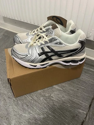 ASICS vita och silver sneakers - Snygga sneakers från ASICS i vitt och silver med svarta detaljer. Skorna har en sportig design med mesh- och syntetmaterial för bra andningsförmåga. Klassisk låg modell med vita snören och bekväm vadderad insida. Perfekta för en trendig streetwear-look.