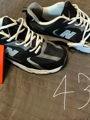 New Balance svarta och grå sneakers - Snygga New Balance sneakers i svart och grått med vita detaljer och snörning. Skorna har en sportig design med mesh och syntetmaterial, samt den klassiska N-loggan på sidan. Perfekta för en streetwear-look eller till vardagsoutfiten.