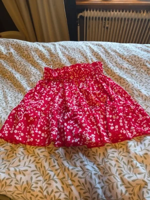 Röd blommig kjol från SHEIN - Söt röd kortkjol från SHEIN med vitt blommigt mönster och bred resår i midjan. Kjolen har volangdetaljer och är tillverkad i ett lätt och luftigt material, perfekt för varma dagar. Endast använd fåtal gånger