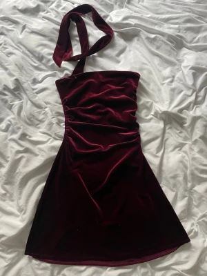 Vinröd sammetsklänning oneshoulder - Vinröd kort klänning i mjuk sammet med oneshoulder-design och bred halsrem. Figurnära passform som markerar midjan och ger en elegant siluett. Perfekt för dig som vill sticka ut med en trendig och lyxig look.