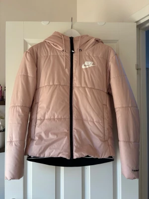 Ljusrosa pufferjacka från Nike - Snygg ljusrosa pufferjacka från Nike med huva och dragkedja framtill. Jackan har Nike-logga på bröstet och är tillverkad i Therm-FIT-material som håller dig varm. Perfekt för kalla dagar och har en modern, sportig look.