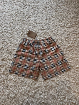 Beige rutiga shorts från Burberry - Säljer ett par klassiska beige shorts från Burberry med det ikoniska rutiga mönstret i svart, vitt och rött. Shortsen har elastisk midja och är perfekta för dig som vill ha en stilren och tidlös look. Materialet känns lätt och passar perfekt till sommaren.