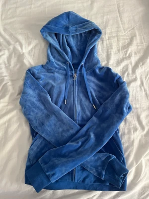 Blå velour hoodie från Juicy Couture - Säljer en blå hoodie från Juicy Couture i mjuk velour med dragkedja framtill och snörning i huvan. Hoodien har långa ärmar, ribbade muddar och ett diskret Juicy Couture-logotyp broderat på bröstet. Perfekt för en chill och trendig look.