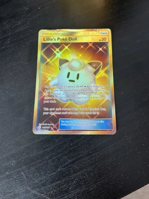 Lillie's Poké Doll Pokémonkort - Säljer ett Lillie's Poké Doll tränarkort från Pokémon TCG. Kortet har en glittrig, holografisk yta med motiv av en söt docka och guldfärgade detaljer. Perfekt för samlare eller dig som vill uppgradera din kortlek.