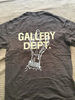 Svart Gallery Dept. t-shirt - Cool svart t-shirt från Gallery Dept. med trycket 'BURIED ALIVE!' på framsidan och ett stort handmotiv samt logga på ryggen. Klassisk rund hals och korta ärmar. Perfekt för dig som gillar streetwear och statement-plagg.